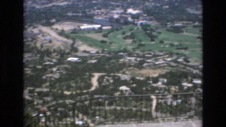 1973 Colorado Springs 8mm frame looking down from an overlook toward forested residential areas, pale roads, green fairways, and sand traps with the railing crossing the lower frame.