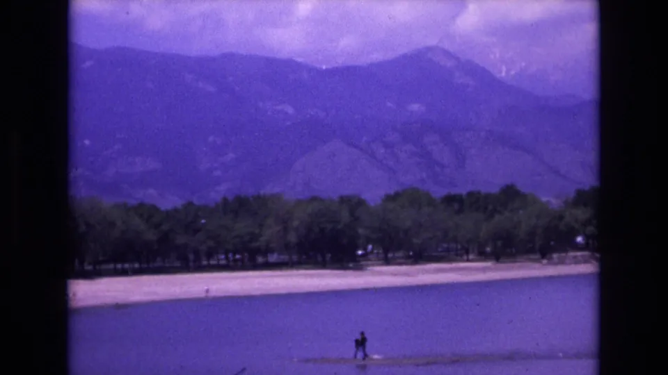 1973 Colorado Springs 8mm frame cutting to a purple-tinted lakeside mountain view with trees along the shore and small distant people near or on the water.