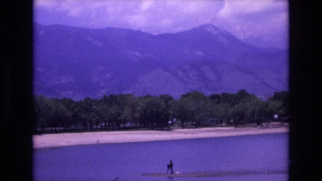 1973 Colorado Springs home-movie frame showing a calm lake, pale shoreline, trees, distant mountains, and a small figure near the water.