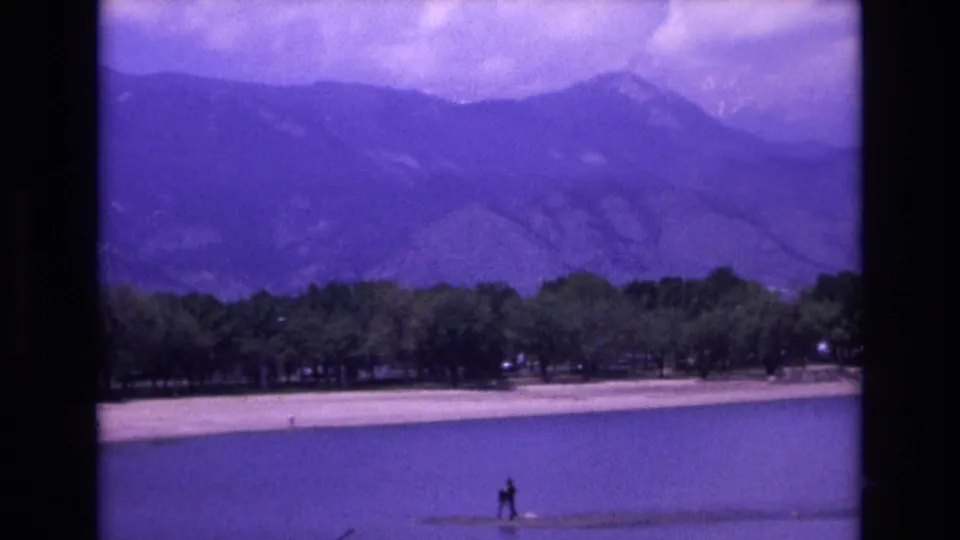 1973 Colorado Springs home-movie frame showing a calm lake, pale shoreline, trees, distant mountains, and a small figure near the water.