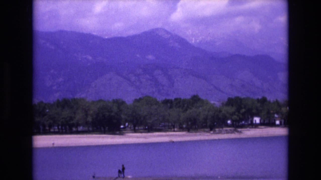 Lake shoreline with trees, mountains, clouds, and small distant figures near the water.