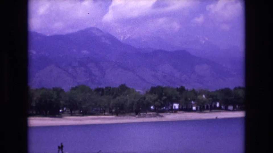 1973 Colorado Springs home-movie view across a lake toward a tree-lined shore, small distant buildings, and a hazy mountain backdrop.