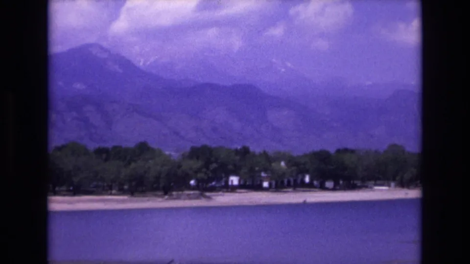 1973 Colorado Springs frame showing calm water, a low shoreline with trees and small buildings, and blue-purple mountain ridges in the background.