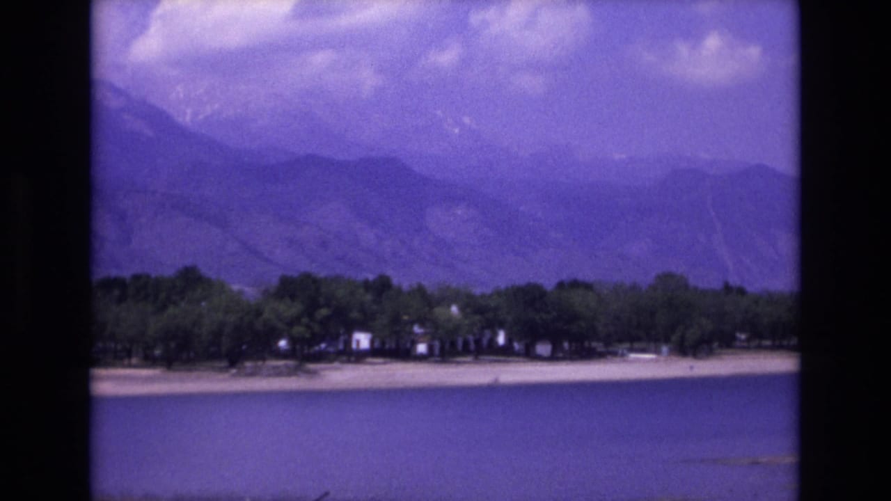 1973 8mm home-movie frame from the Colorado Springs area showing blue lake water, a pale beach, trees, small shore structures, and hazy mountain ridges.
