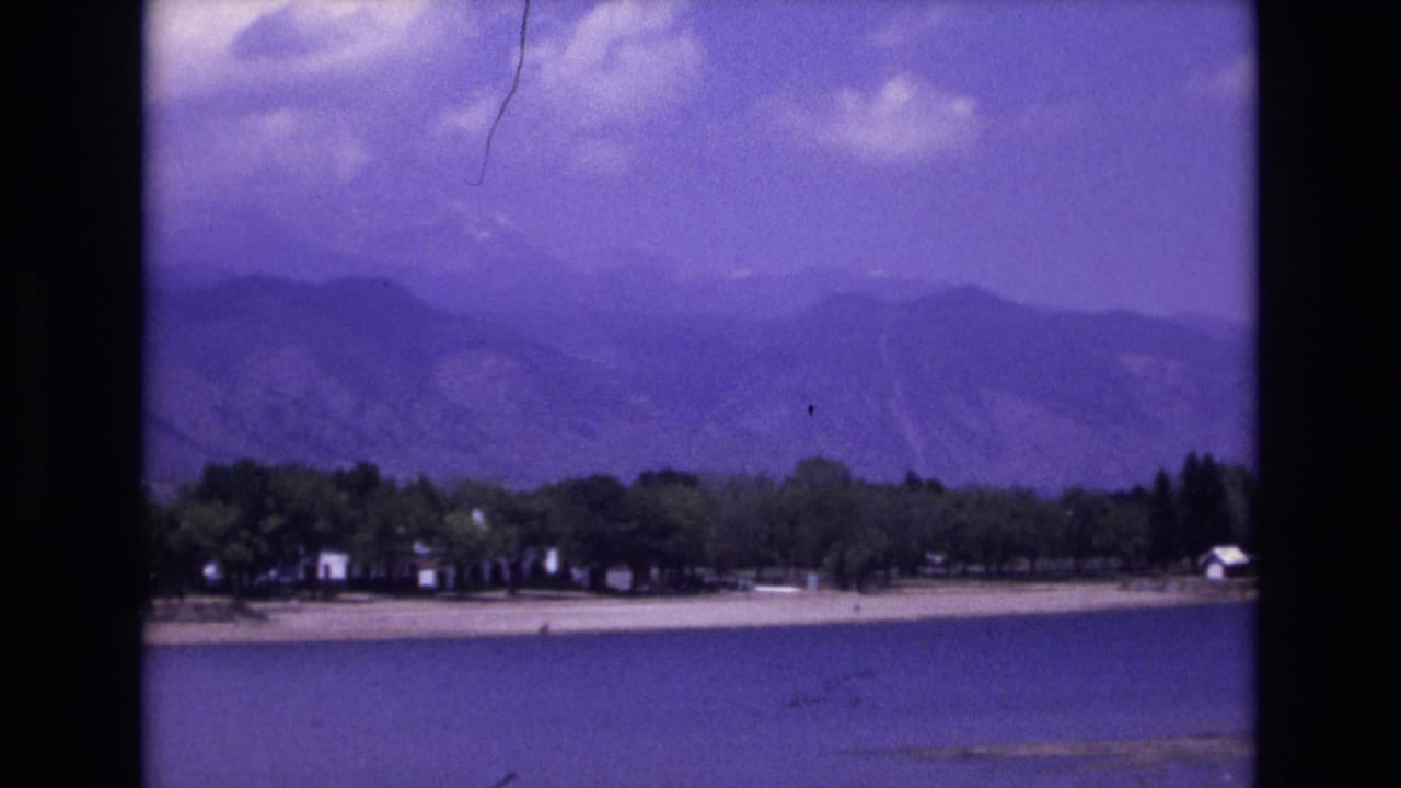 1973 Colorado Springs area lake view with a sandy shoreline, scattered trees, small structures, distant mountains, clouds, and visible 8mm film scratches.