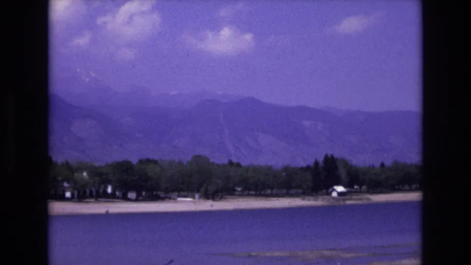 1973 8mm frame showing a wide Colorado Springs area lakeside vista with water in the foreground, a pale beach, trees, a small white structure, and layered mountains beyond.