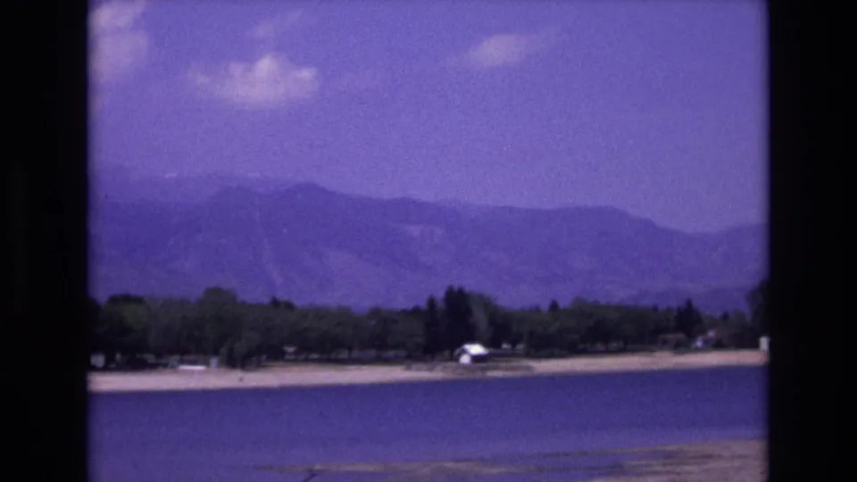 Final 1973 Colorado Springs area frame holding on blue lake water, a low shoreline, scattered trees, a small white lakeside structure, and hazy mountain ridges under a purple-tinted sky.