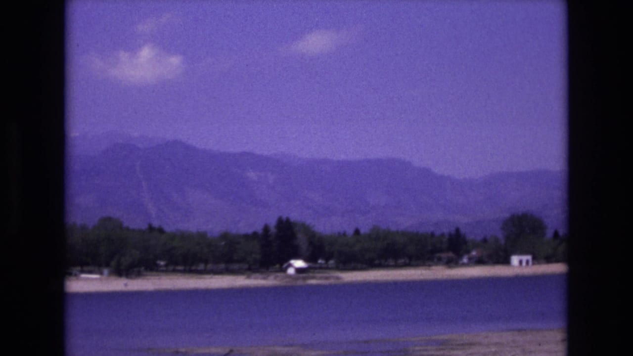 1973 8mm home-movie view near Colorado Springs showing a calm lake, sandy shoreline, trees, small white structures, and hazy mountain ridges.