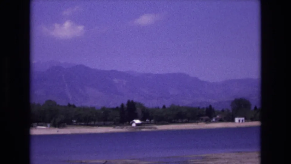 1973 8mm home-movie view near Colorado Springs showing a calm lake, sandy shoreline, trees, small white structures, and hazy mountain ridges.