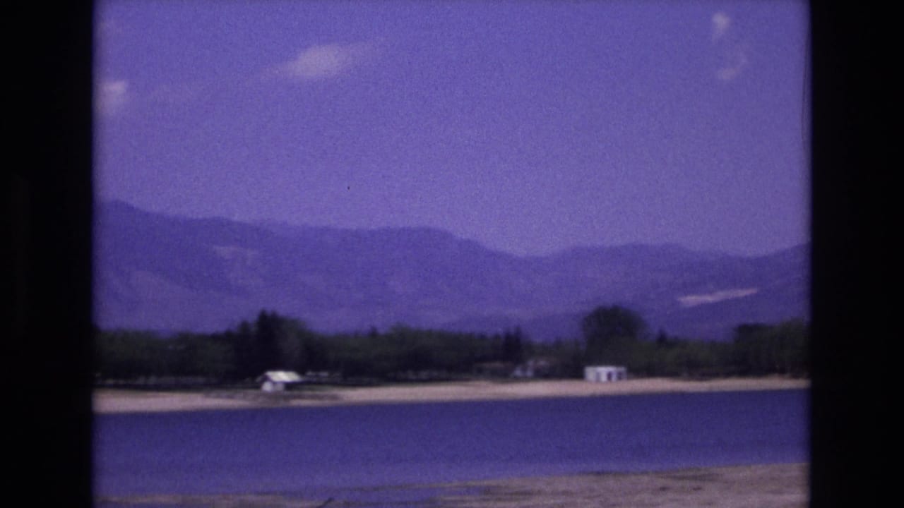 Colorado Springs-area lakeside landscape from 1973 with blue water in the foreground, a tree-lined shore, small white buildings, and soft mountain ridges beyond.