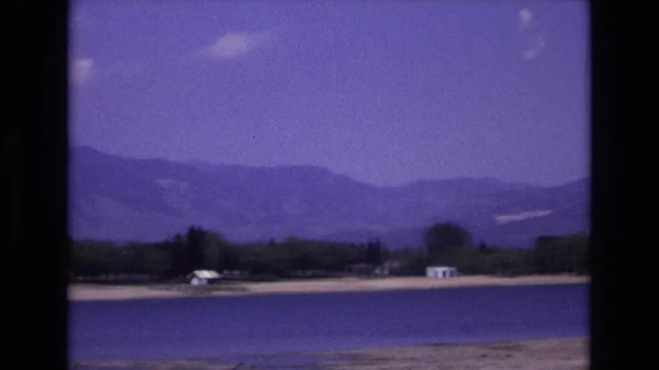 Colorado Springs-area lakeside landscape from 1973 with blue water in the foreground, a tree-lined shore, small white buildings, and soft mountain ridges beyond.