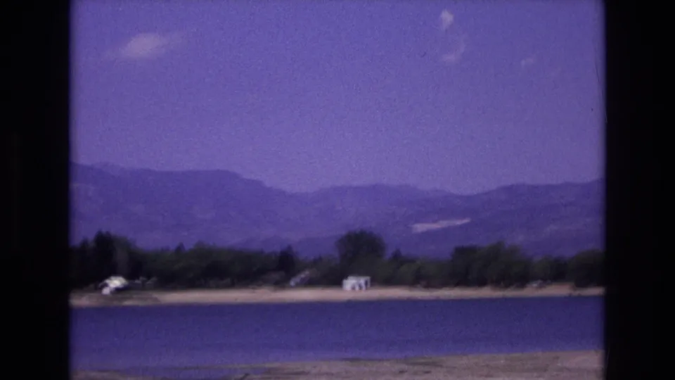 Soft 8mm frame of a Colorado Springs lakeshore in 1973, with water, sandy ground, shoreline trees, a small white structure, and hazy mountains behind.