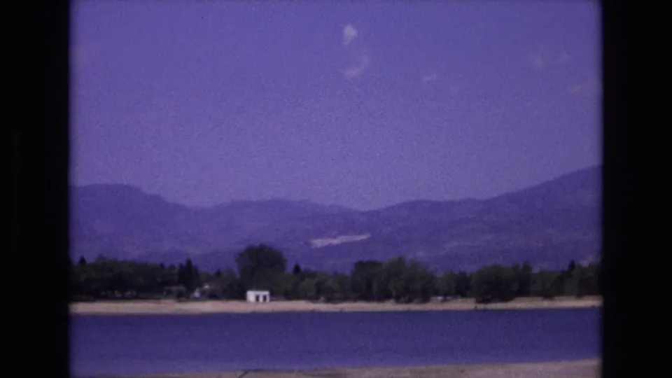 Wide 1973 Colorado Springs-area lake view with a low sandy shore, dark tree line, small white building, hazy mountain backdrop, and pale clouds in a purple-tinged sky.