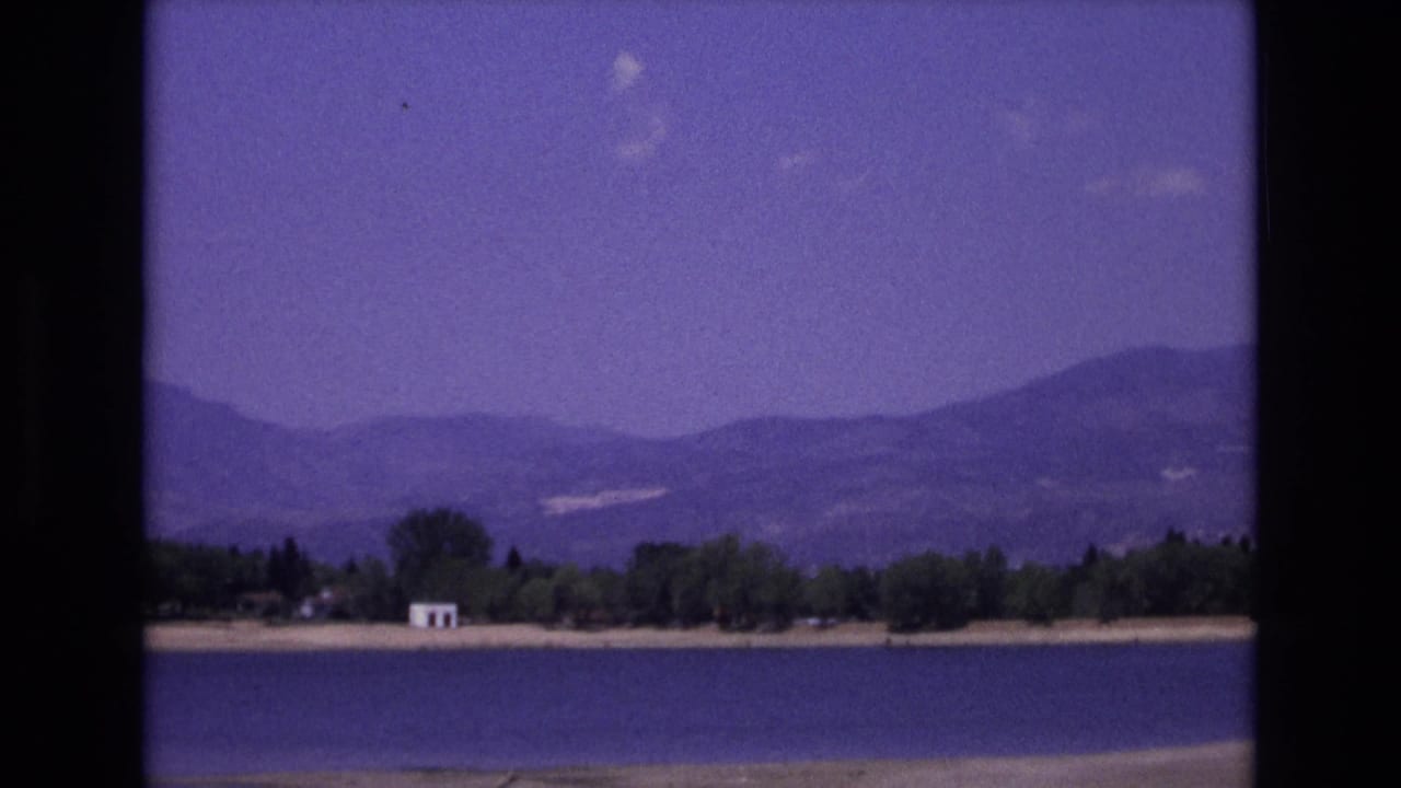 1973 Colorado Springs 8mm frame showing a lake, tree-lined shore, distant mountains, and a small white shoreline structure under a color-shifted sky.