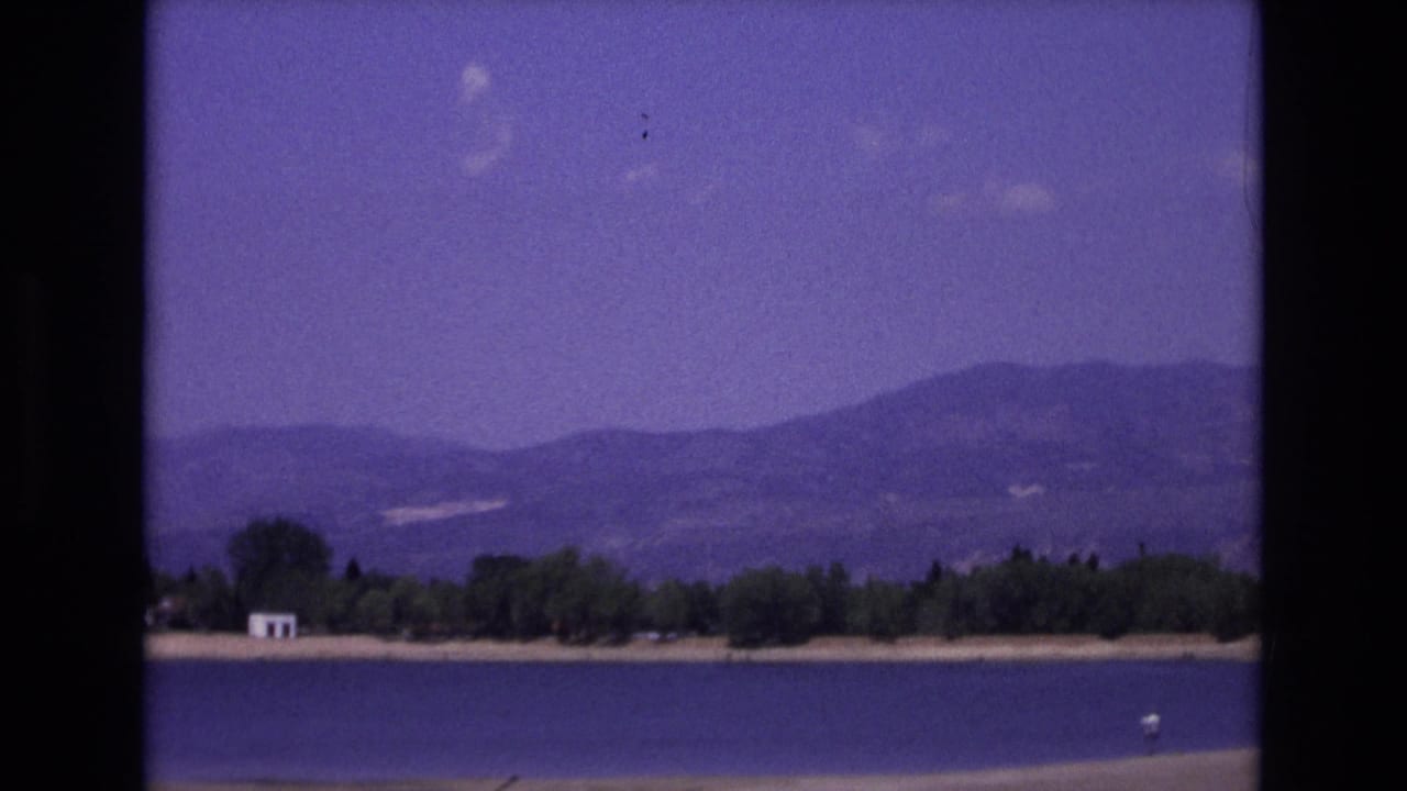 1973 Colorado Springs home-movie frame of a lake before distant mountains, with trees, a small white structure, and a small light object on the water.