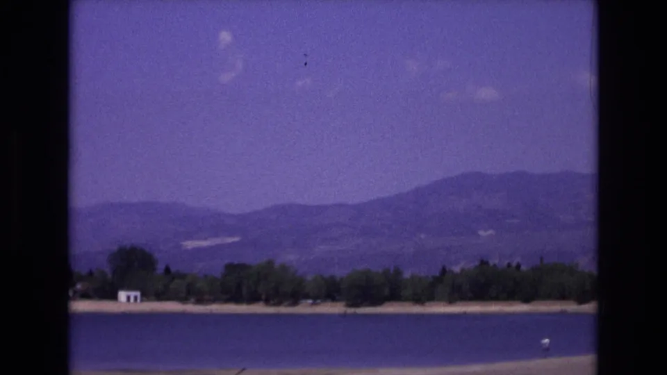 1973 Colorado Springs home-movie frame of a lake before distant mountains, with trees, a small white structure, and a small light object on the water.
