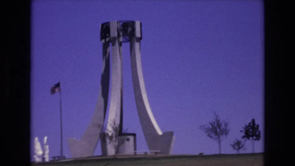 1973 Colorado Springs 8mm frame showing a mid-century concrete monument with curved vertical supports, an American flag, young trees, and open sky.