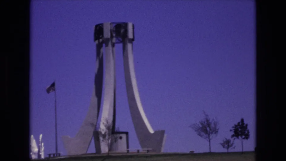1973 Colorado Springs home-movie frame of a concrete public monument with curved supports, a nearby American flag, small trees, and a grassy foreground.