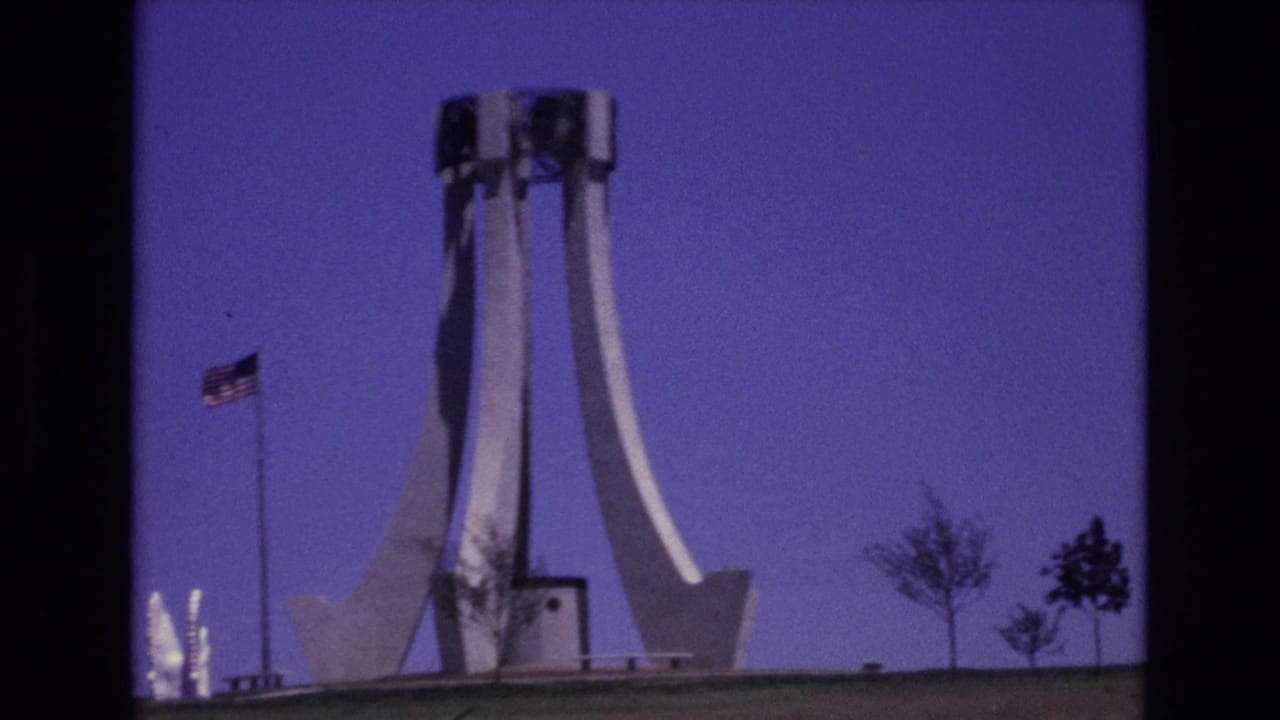 1973 Colorado Springs home-movie frame showing a white modernist monument, an American flag on a pole, sparse trees, and a deep blue sky.