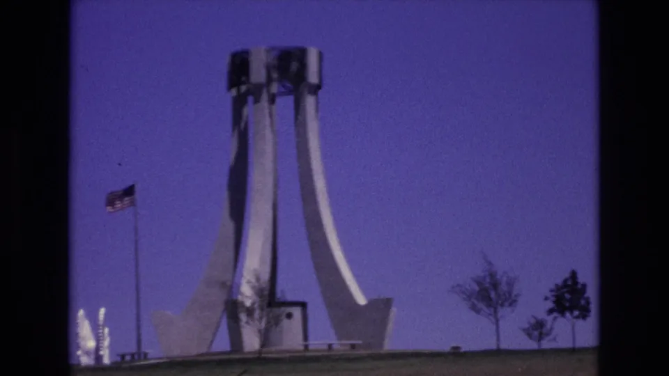 1973 Colorado Springs home-movie frame showing a white modernist monument, an American flag on a pole, sparse trees, and a deep blue sky.