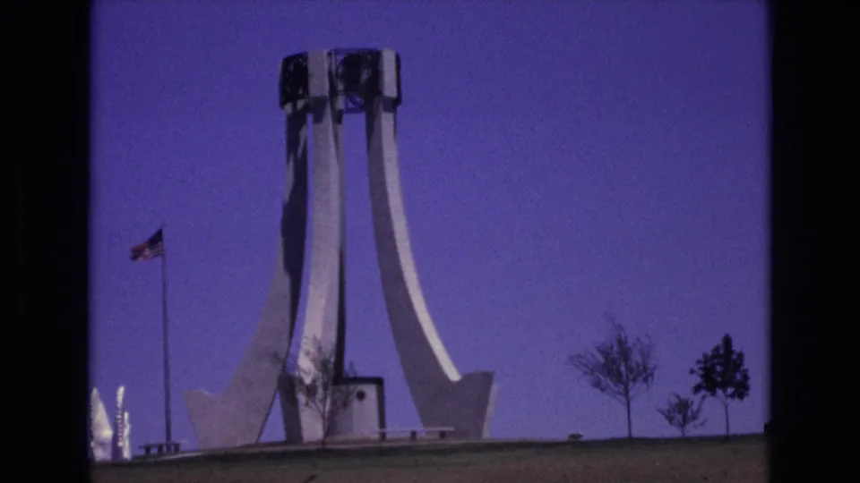 1973 Colorado Springs frame with the flagpole left of a white monument, landscaped grass in front, and small trees along the horizon.