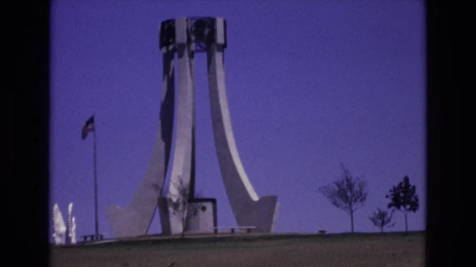 1973 Colorado Springs home-movie still showing a flag waving beside a tall white monument, with sparse trees and saturated 8mm color.