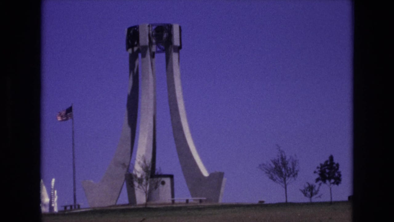 1973 Colorado Springs 8mm frame showing a white modernist monument on a lawn, an American flag on a pole, sparse trees, and a deep purple sky.