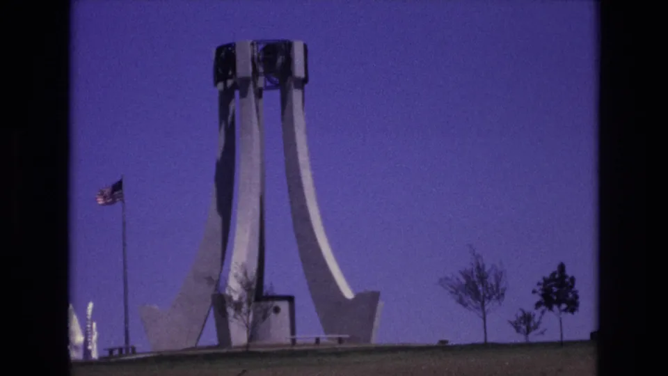 1973 Colorado Springs 8mm frame showing a white modernist monument on a lawn, an American flag on a pole, sparse trees, and a deep purple sky.