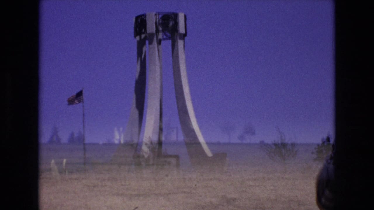 1973 Colorado Springs 8mm frame with a distant white sculptural monument, American flag, open grass, faint haze, and soft purple-blue color cast.