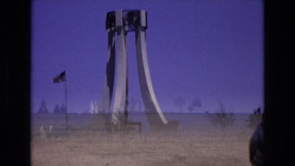1973 Colorado Springs 8mm frame with a distant white sculptural monument, American flag, open grass, faint haze, and soft purple-blue color cast.