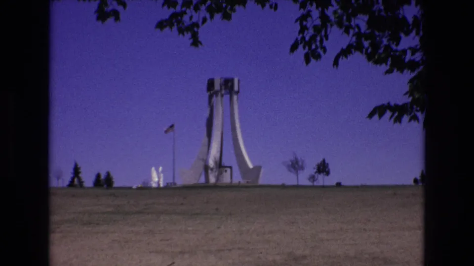 Wide 1973 Colorado Springs 8mm view of the modernist monument across a grassy field, with an American flag, small trees, and dark branches framing the sky.