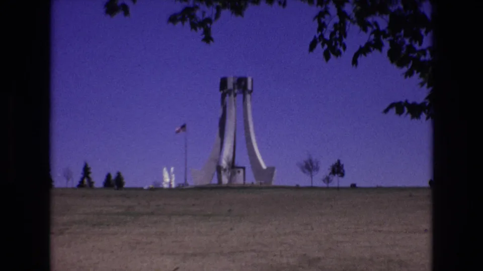 1973 Colorado Springs 8mm frame showing the white modernist monument and American flag from a distance, framed by silhouetted branches over a broad grassy foreground.