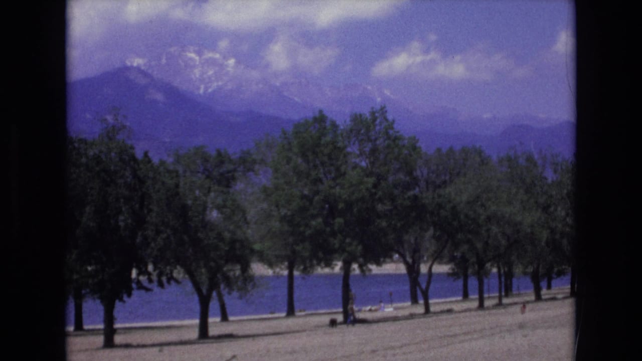 1973 Colorado Springs 8mm frame showing trees beside blue water with snow-dusted mountains in the distance and small shoreline figures barely visible.