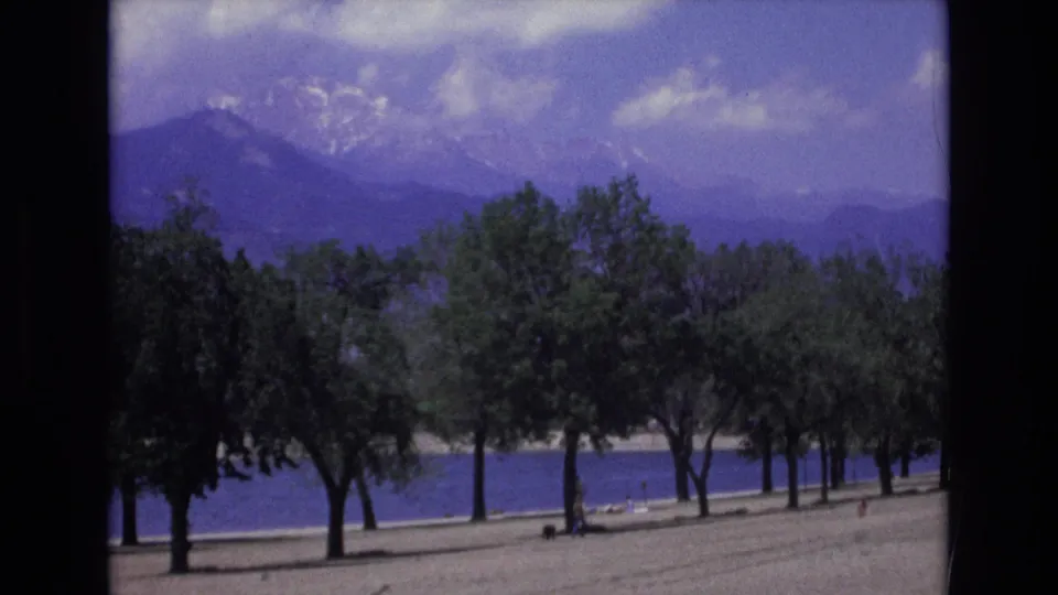 1973 Colorado Springs 8mm frame showing trees beside blue water with snow-dusted mountains in the distance and small shoreline figures barely visible.