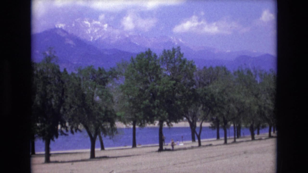 Tree-lined shoreline beside blue water with mountains and clouds in the background.