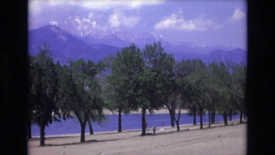 1973 Colorado Springs 8mm frame with a row of trees on the shore, calm blue water behind them, and hazy snow-capped mountains beyond.