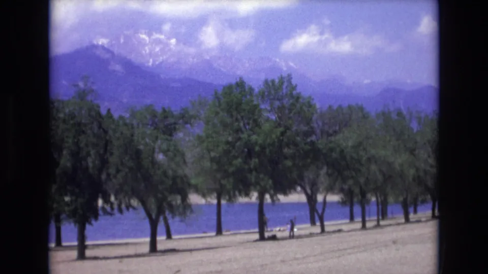 1973 Colorado Springs home-movie frame showing a lakeside row of trees, a blue band of water, and distant mountains softened by grain and color shift.