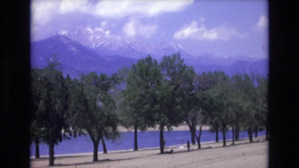 1973 Colorado Springs 8mm frame holding on shoreline trees, blue water, small distant visitors, and a snow-capped mountain backdrop under a partly cloudy sky.