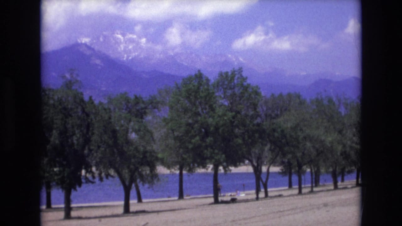 1973 8mm Colorado Springs footage showing a sandy lakeshore, rows of green trees, blue water, and hazy snow-marked Rocky Mountains under scattered clouds.