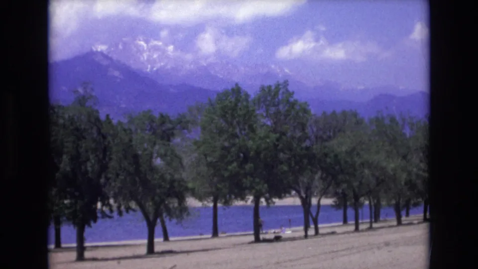 1973 8mm Colorado Springs footage showing a sandy lakeshore, rows of green trees, blue water, and hazy snow-marked Rocky Mountains under scattered clouds.