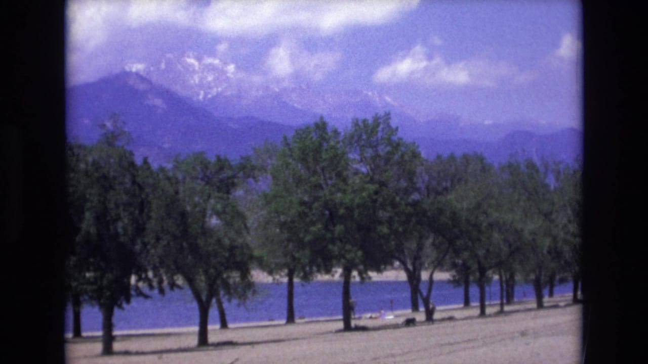 Lakeside trees and sandy shore with mountains and clouds behind them.