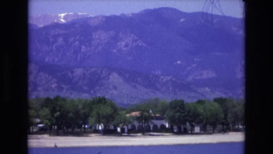 The frame shows Colorado Springs lakeside buildings and parked cars partly screened by trees, with blue water in the foreground and hazy mountains rising behind.