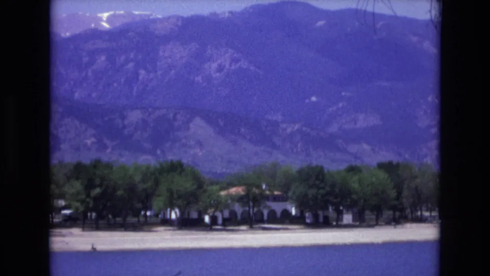 A distant 1973 Colorado Springs waterfront view includes blue lake water, pale sand, tree-shaded low buildings, small cars, and purple-cast Rocky Mountain slopes in the background.