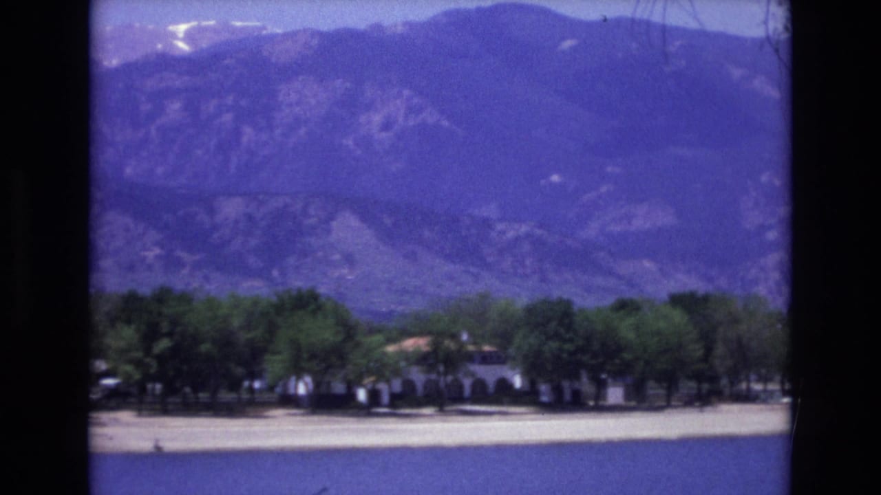 1973 Colorado Springs 8mm frame showing distant mountains above green trees and a low light-colored building near a road or water edge.