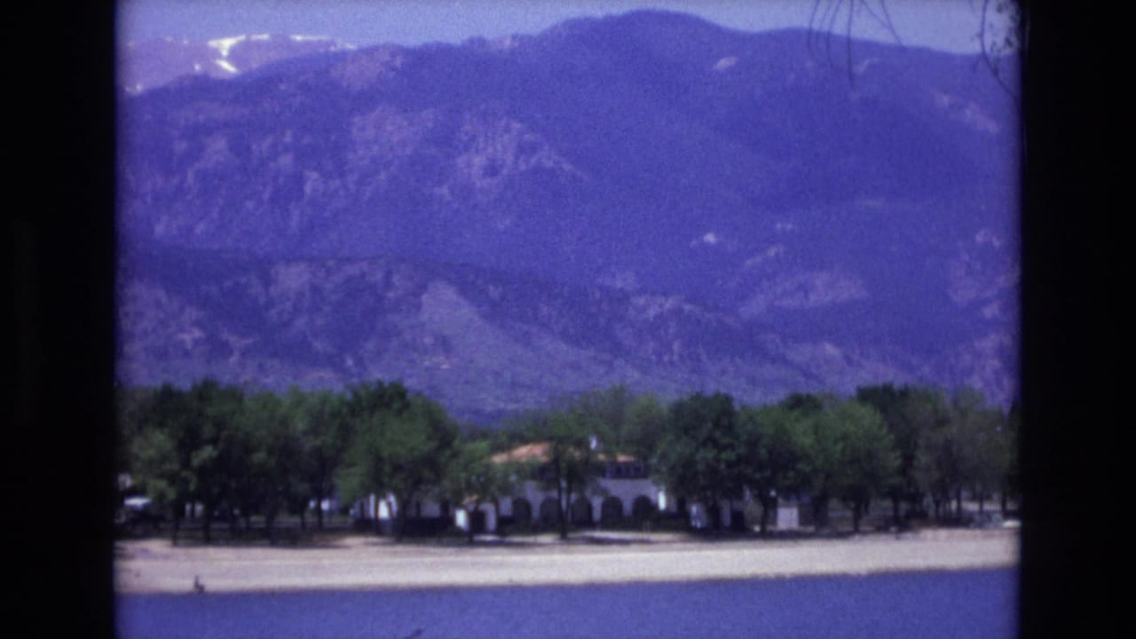 Colorado Springs travel footage frame with a low building partly hidden by trees and a broad mountain backdrop in soft 8mm color.