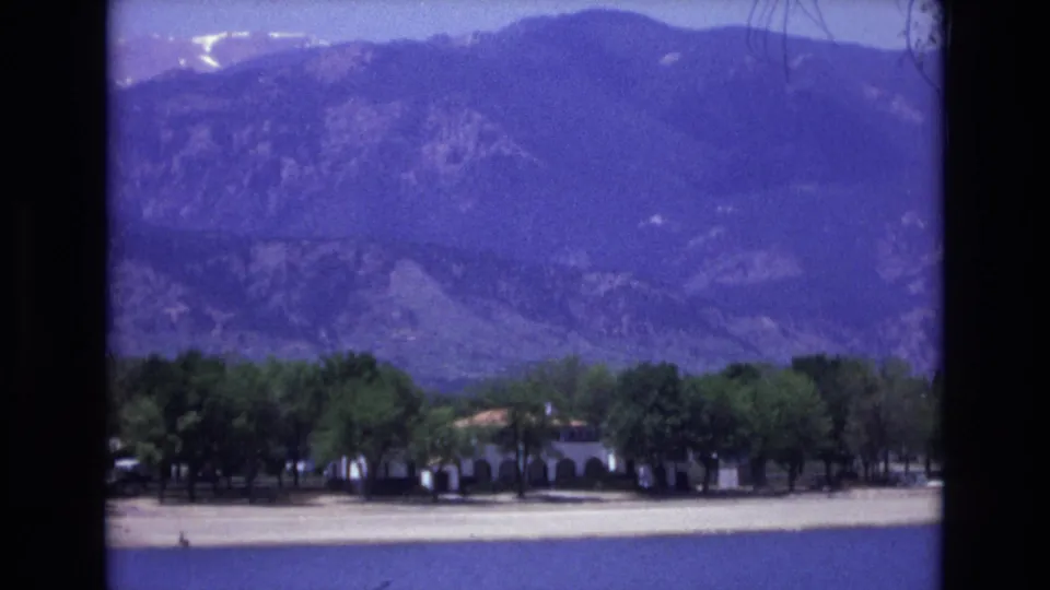 Colorado Springs travel footage frame with a low building partly hidden by trees and a broad mountain backdrop in soft 8mm color.