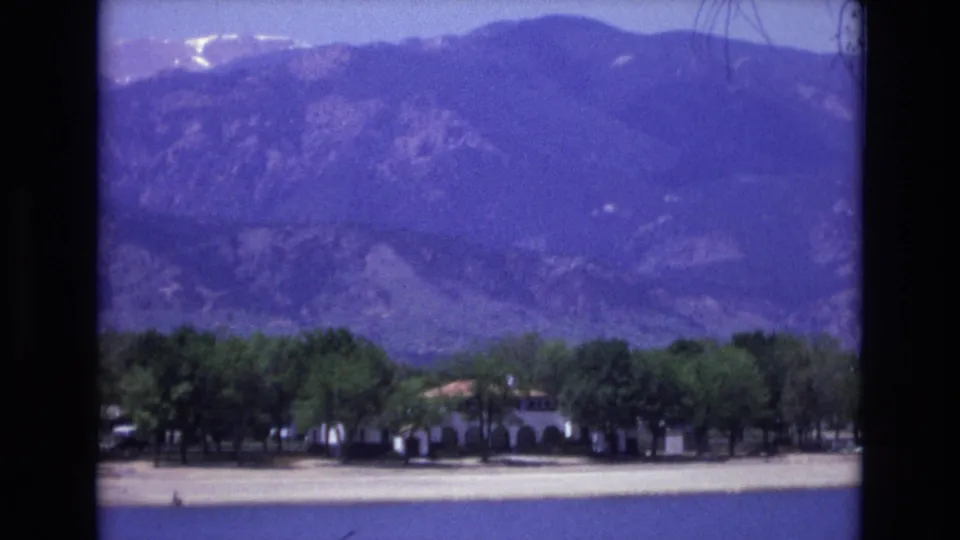 1973 Colorado Springs home-movie frame showing a distant mountain slope, trees, and a low roadside or resort-like building rendered with blue-violet film shift.