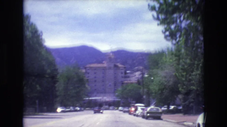 Street-level 1973 Colorado Springs frame showing cars along a road, trees on both sides, mountains in the distance, and a large stone building ahead.