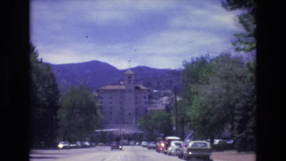 1973 Colorado Springs home-movie frame showing a forward road view with parked cars, a large towered building, trees, and mountain ridges beyond.