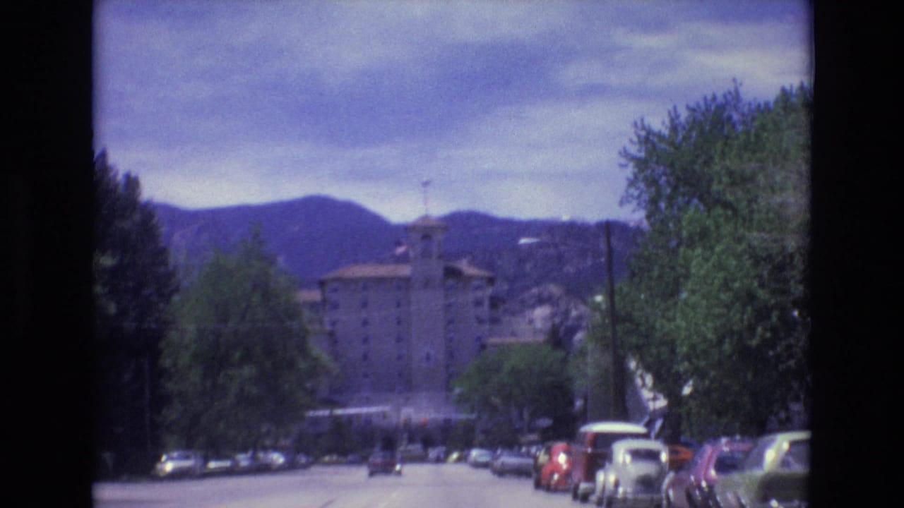 Roadside cars and trees leading toward a large building below mountains.